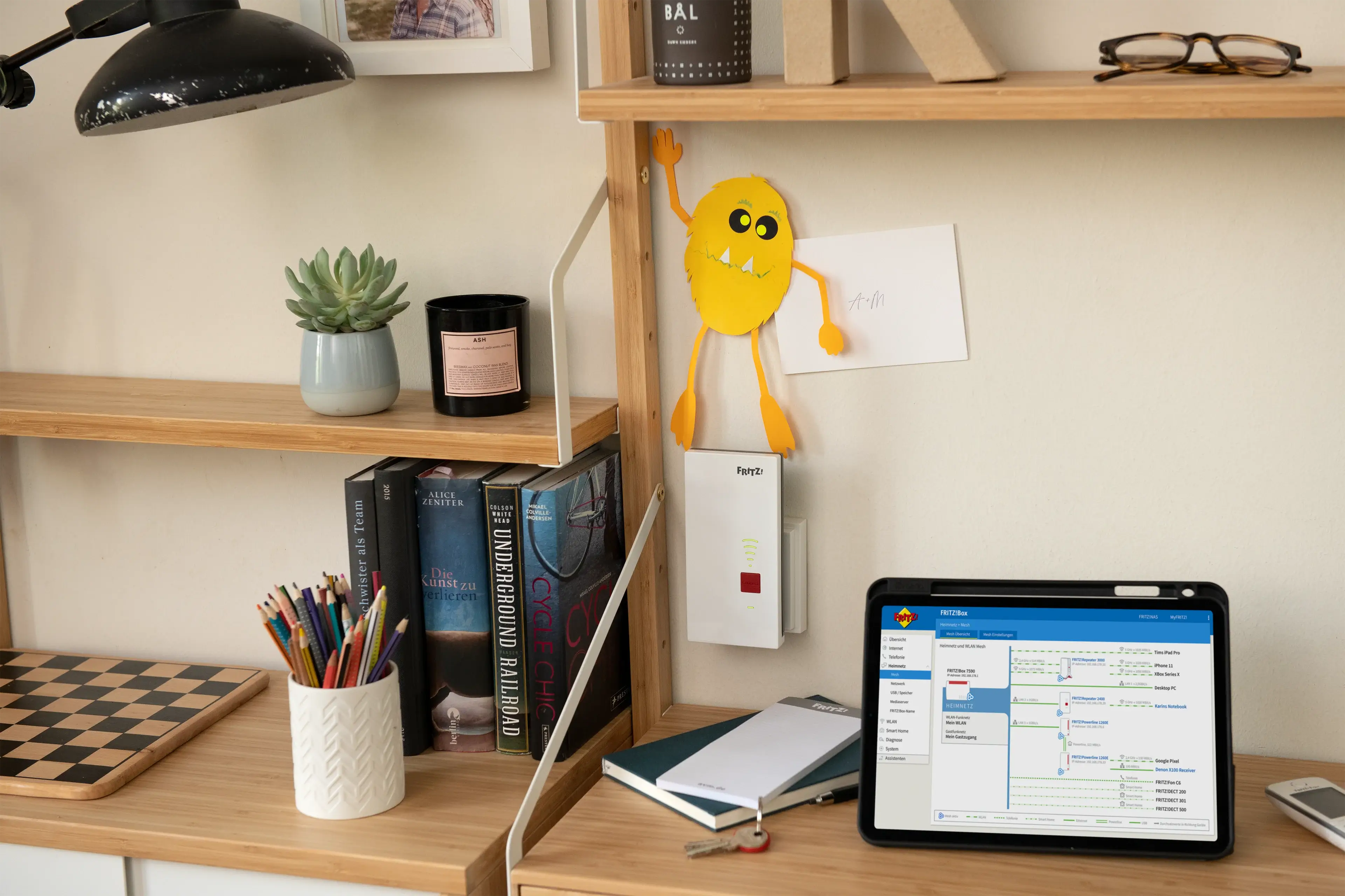 Büro mit Regalen, Schreibtischlampen, einem Tablet und bunten Stiften, dekoriert mit einem gelben Monster aus Papier.